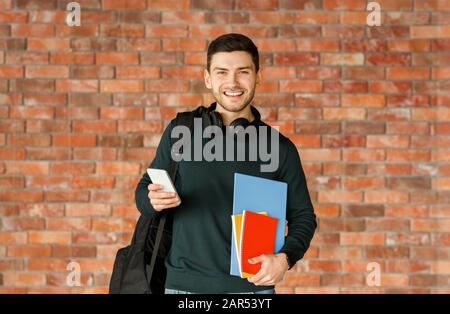 Guy With Backpack Holding Smartphone Standing Over Brick Wall Indoor Stock Photo