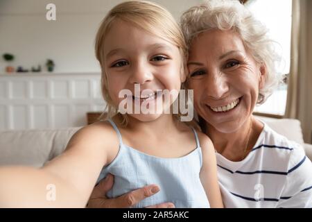 Small preschool granddaughter recording funny video with excited elderly grandmother. Stock Photo