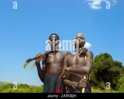 Portrait of two young Mursi warriors holding a stick. The Mursi (or Murzu) are a Sub-Saharan African nomadic cattle herder tribe located in the Omo va Stock Photo