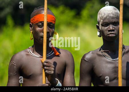 Portrait of two young Mursi warriors holding a stick. The Mursi (or Murzu) are a Sub-Saharan African nomadic cattle herder tribe located in the Omo va Stock Photo