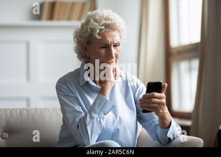 Thoughtful middle aged hoary woman holding smartphone, reading news. Stock Photo