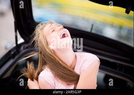 the girl sits in the car on a summer day and laughs out loud, squinting her eyes. Warm positive weekend Stock Photo