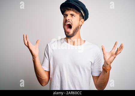 Young driver man with beard wearing hat standing over isolated white background crazy and mad shouting and yelling with aggressive expression and arms Stock Photo