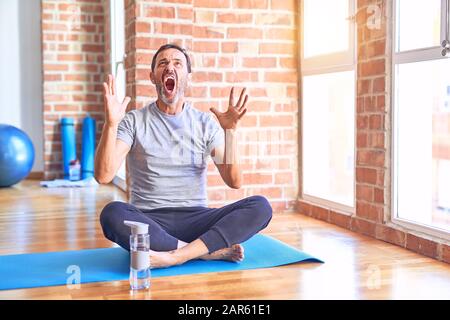 Middle age handsome sportman sitting on mat doing stretching yoga exercise at gym crazy and mad shouting and yelling with aggressive expression and ar Stock Photo