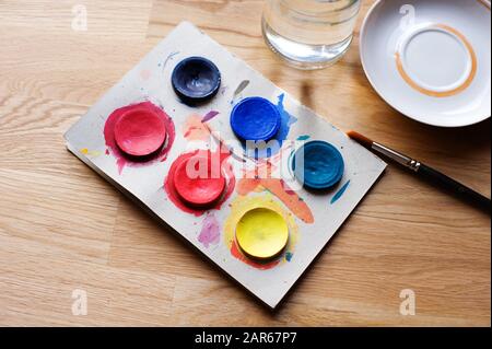 Watercolor paints with brush, plate and glass jar on the wooden table. Stock Photo