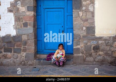 Peru, Cusco 2019-11-29. A little girl in a national dress sits with a baby llama on the steps in front of the door. Concept of hard life of children Stock Photo