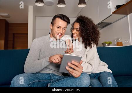 Romantic Date. Young multiethnic couple at home sitting on sofa browsing digital tablet together smiling cheerful Stock Photo