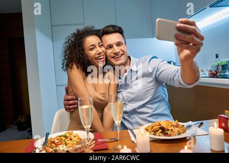 Romantic Date. Young multiethnic couple at home having dinner hugging taking selfie on smartphone saving moment laughing cheerful Stock Photo