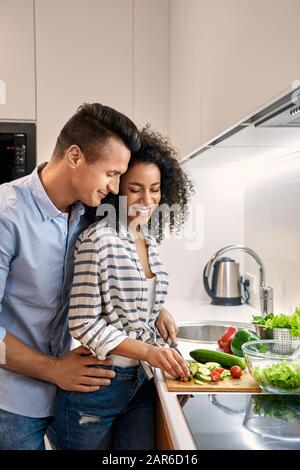 Romantic Date. Young multiethnic couple standing at kitchen cooking dinner husband hugging wife cutting vegetables smiling cheerful Stock Photo