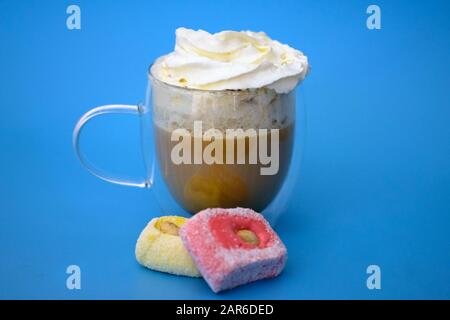 Cappuccino with whipped cream on a blue background. Double bottom cup with coffee and pink rahat lakoum candies. Tasty hot drink in a transparent cup. Stock Photo