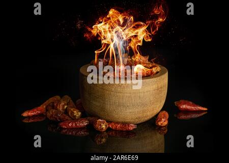 Lot of small hot peppers with burning fire in wooden bowl isolated on black glass Stock Photo