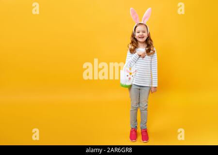 A little girl is standing and holding a bag for Easter eggs and sweets. Happy child in full-growth in bunny ears on a yellow background. Stock Photo