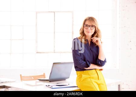 Portrait shot of elegant mature businesswoman looking at camera and smiling while standing at office desk. Stock Photo