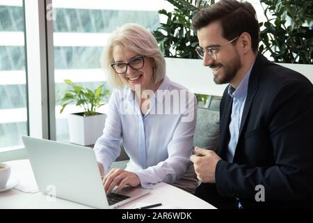 Happy middle aged leader working on computer with young colleague. Stock Photo