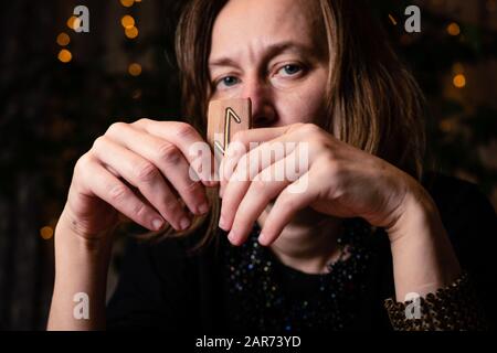 Fortune telling on the runes. A mature female witch holds a rune near her face, meaning 'world tree', 'protection'. Close-up portrait. Stock Photo