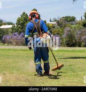 Man wearing protective clothing and safety helmet strimming grass in a garden. Western Cape, South Africa Stock Photo