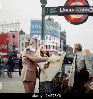 London 1970s, elegant couple reading newspaper, underground public subway exit, tube sign, evening, Piccadilly Circus, England, UK, GB, Great Britain, Stock Photo