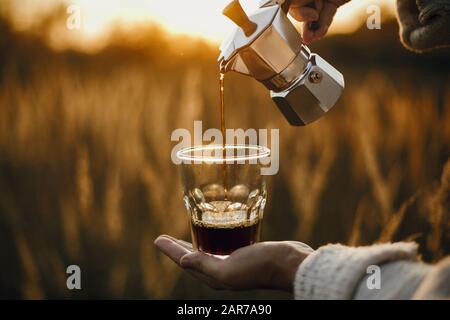 Traveler pouring fresh hot coffee from geyser coffee maker into glass cup in sunny warm light in rural countryside herbs. Atmospheric tranquil moment. Stock Photo