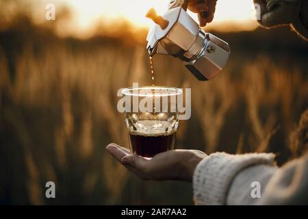 Traveler pouring fresh hot coffee from geyser coffee maker into glass cup in sunny warm light in rural countryside herbs. Atmospheric tranquil moment. Stock Photo