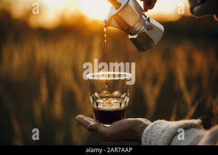 Hipster pouring fresh hot coffee from geyser coffee maker into glass cup in sunny warm light in rural countryside herbs. Atmospheric tranquil moment. Stock Photo