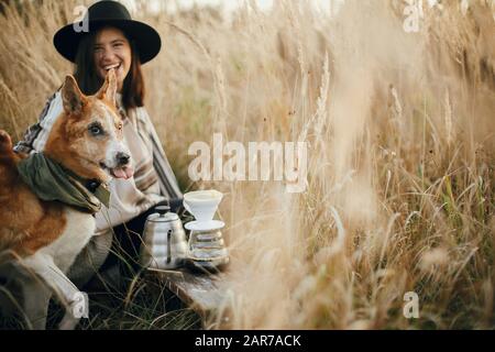 Stylish hipster traveler smiling with her funny cute dog and  brewing fresh coffee in rural countryside herbs in sunset light. Atmospheric rustic mome Stock Photo
