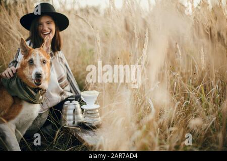 Stylish hipster traveler hugging her golden dog and smiling, brewing fresh coffee in rural countryside herbs in sunset light. Atmospheric rustic momen Stock Photo