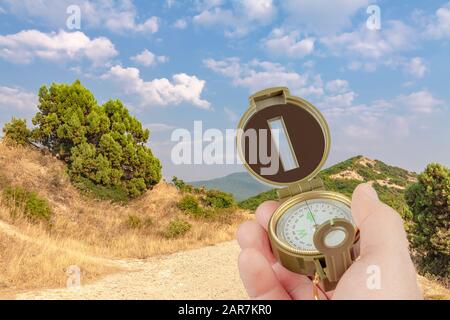 A hand holds a compass on a background of mountains, trees, bushes and sky. Stock Photo