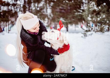 Young girl hugs dog with decorative deer horns Christmas labrador retriever in winter forest Stock Photo