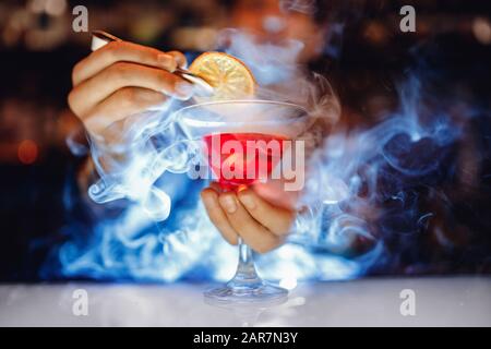 Barman is preparing red alcoholic cocktail with smoke in martini glass, decorated with orange Stock Photo