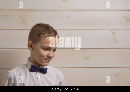 A fair-haired boy of 10 years in a white shirt with the self-tie bow tie smiles against the background of a wooden wall Stock Photo