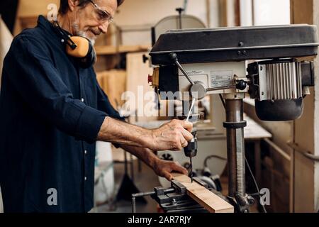 Mature worker in carpentry preparing furniture parts using a drilling machine Stock Photo