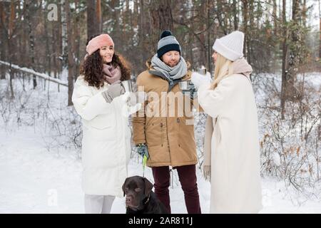Happy young man with black retriever and two girls having hot tea and chatting Stock Photo