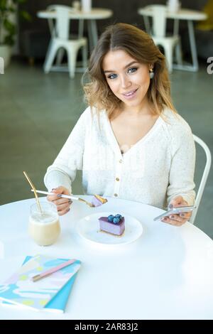 Young cheerful woman using smartphone and having blueberry cheesecake Stock Photo