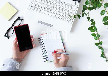 Modern workplace in white color with woman hand using mobile phone and making some notes in a notebook. Top view. Flat lay. Stock Photo