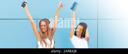 Fashion portrait of two pretty smiling hipsters woman in sunglasses holding smartphone and dancing against the colorful blue wall. Stock Photo