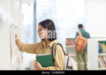 Side view portrait of female Asian student wearing glasses choosing books on shelves in college library, copy space Stock Photo