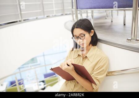 High angle portrait of young Asian woman writing in notebook while standing by spiral staircase in modern college interior, copy space Stock Photo