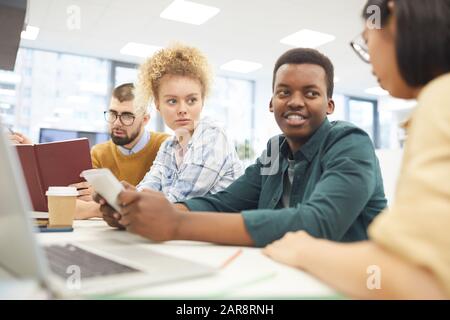 Multi-ethnic group of students studying in college library, focus on African-American man smiling at partners Stock Photo
