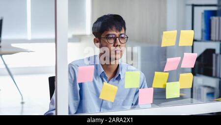 asian businessman looking and writing on sticky note for brainstorming ideas on clear whtieboard at modern office. analyze and synthesis Stock Photo