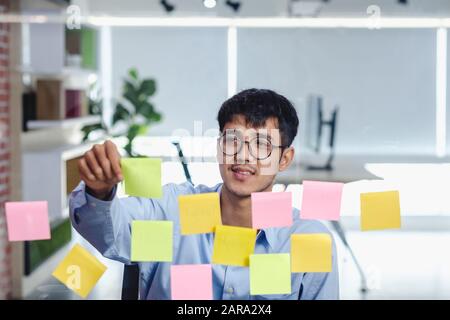 asian businessman looking and writing on sticky note for brainstorming ideas on clear whtieboard at modern office. analyze and synthesis Stock Photo