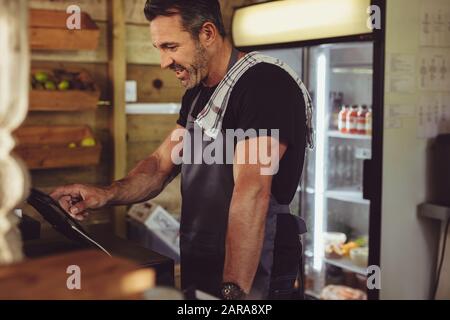 Male barista at counter using cashbox computer in cafe store checking client's order. Waiter in apron working in coffee shop. Stock Photo