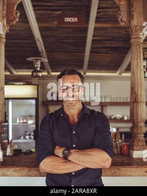 Portrait of a smiling cafe owner standing with his arms crossed. Handsome coffee shop owner looking at camera and smiling. Stock Photo