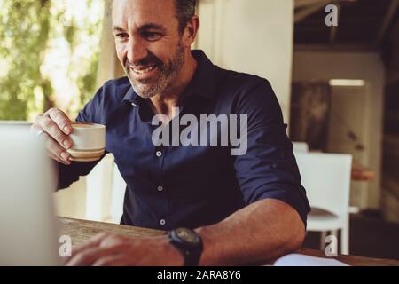 Smiling businessman using laptop and having coffee while sitting at table in coffee shop. Man drinking a cup of coffee and looking at his laptop compu Stock Photo