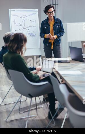 Smiling businesswoman standing in front of flip chart for presentation with team. Woman giving presentation on sales strategies to colleagues in board Stock Photo