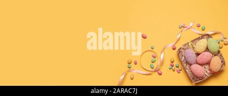Happy easter composition. Decor eggs in a wooden wicker basket and sweet candy on a yellow background. Can be used for web banner, poster, voucher. Stock Photo