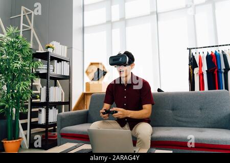 Amazed Asian man in casual clothes with open mouth playing video games using VR glasses and gamepad sitting on sofa in cozy room Stock Photo