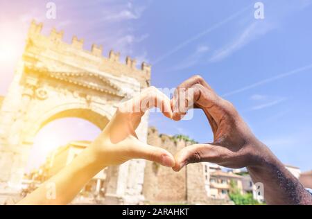 Closeup of woman and man hands showing heart shape during romantic vacation - Young multiracial couple making love symbol next a roman monument - Mult Stock Photo