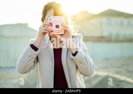Young woman shooting with pink instant camera - Trendy girl taking snapshots outdoor with back sun light - New generation trends concept - Focus on sh Stock Photo