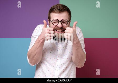 Funny bearded man in round glasses, makes an approving gesture with hands, thumbs up, closes eyes and grins teeth. Stock Photo