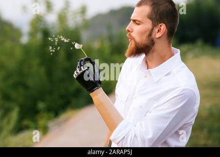 Man holding dandelion in a bionic hand, seeds fly as he blowing on it. Side view Stock Photo